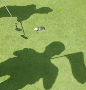 shadows of two golfers on putting green at hole with ball on edge of cup